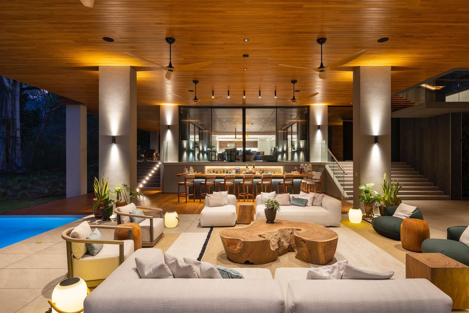 An evening room with warm timber ceilings and an illuminated bar, opening toward the pool.