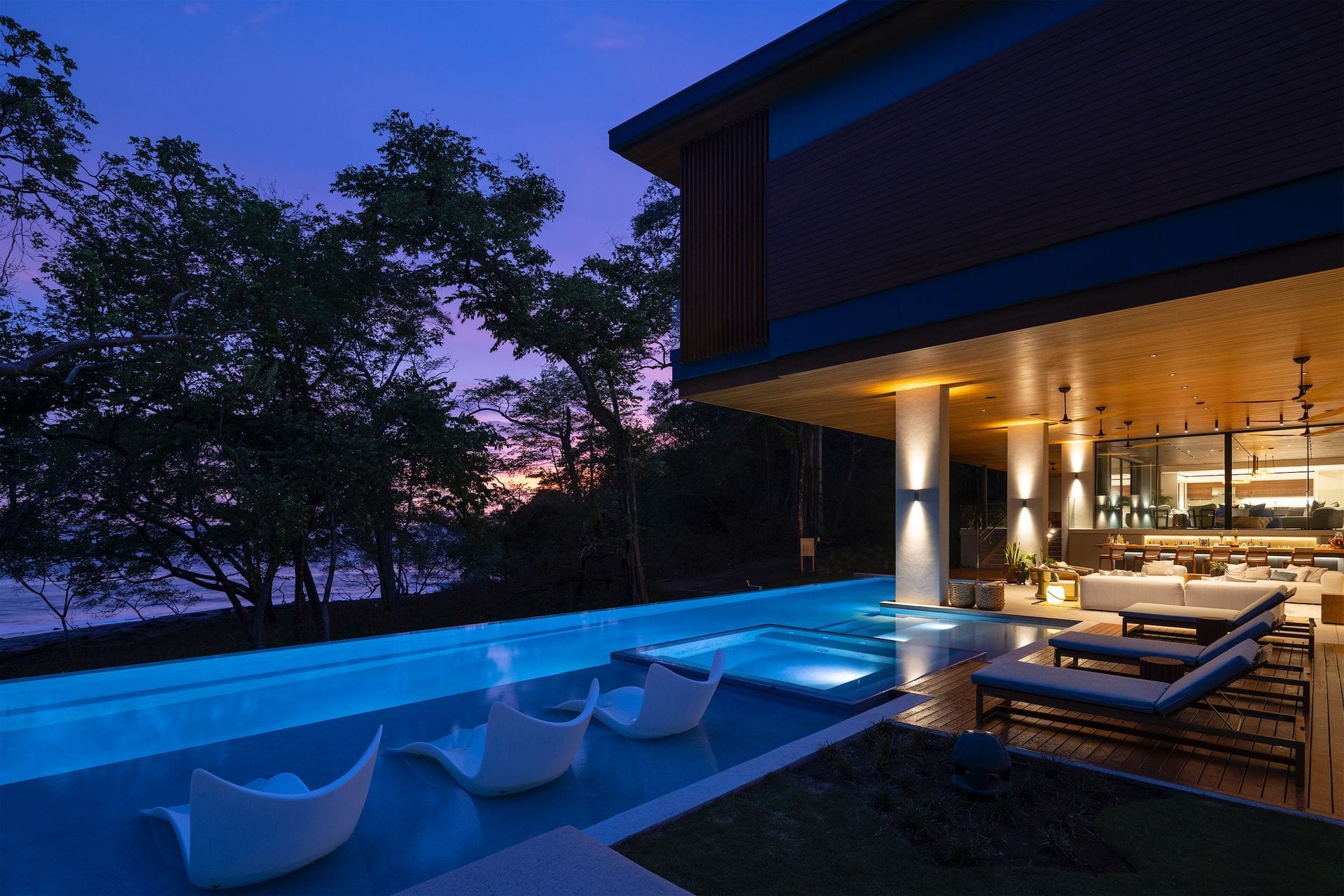Illuminated columns and warm rooms stand beside a blue pool at dusk.